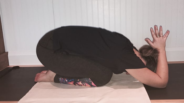 moça de cabelo preto e roupa preta, ajoelhada, com bumbum apoiado nos calcanhares, tronco para frente, testa relaxada no solo, cotovrlos apoiados no solo ao lado do corpo, mãos unidas acima da cabeça