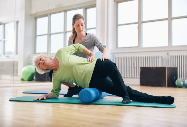 mulher idosa de calça preta e blusa verde deitada de lado em postura de Pilates em cima do rolinho com instrutora atrás auxiliando